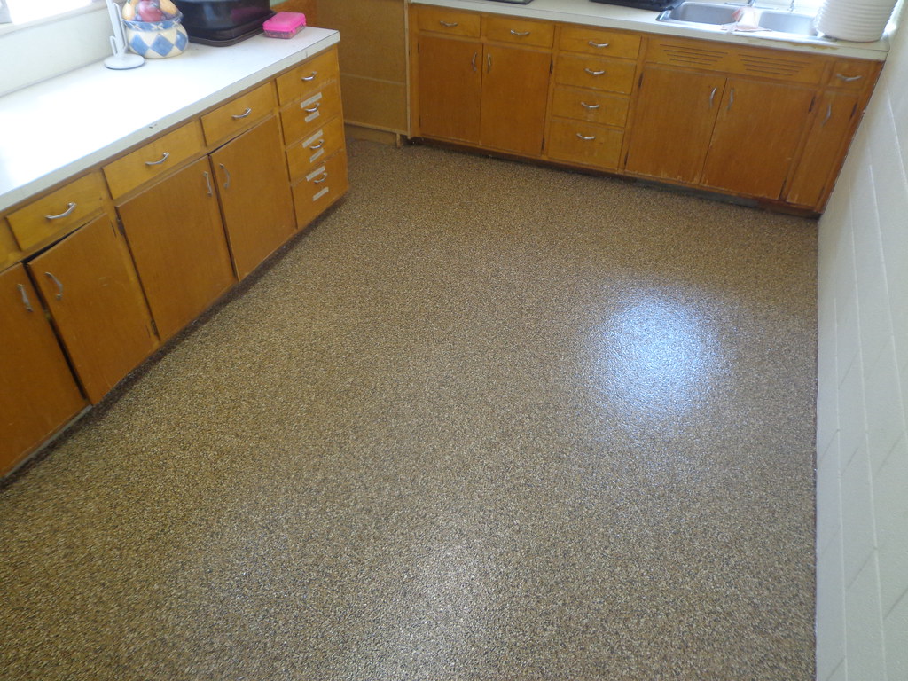 Tan and brown flake epoxy floor in a small kitchen with wood cabinets and white countertops, daylight from a window reflecting off the clear coat in Carrollton, TX